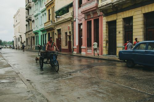 Free Man rides tricycle on a wet street in an urban area with colorful buildings and classic car. Stock Photo