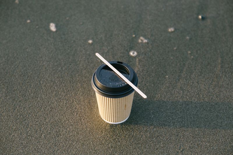 A disposable coffee cup with a lid and stirrer placed on a sandy beach under daylight.