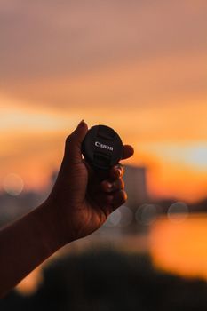 A close-up of a hand holding a camera lens cap against a vibrant sunset background.