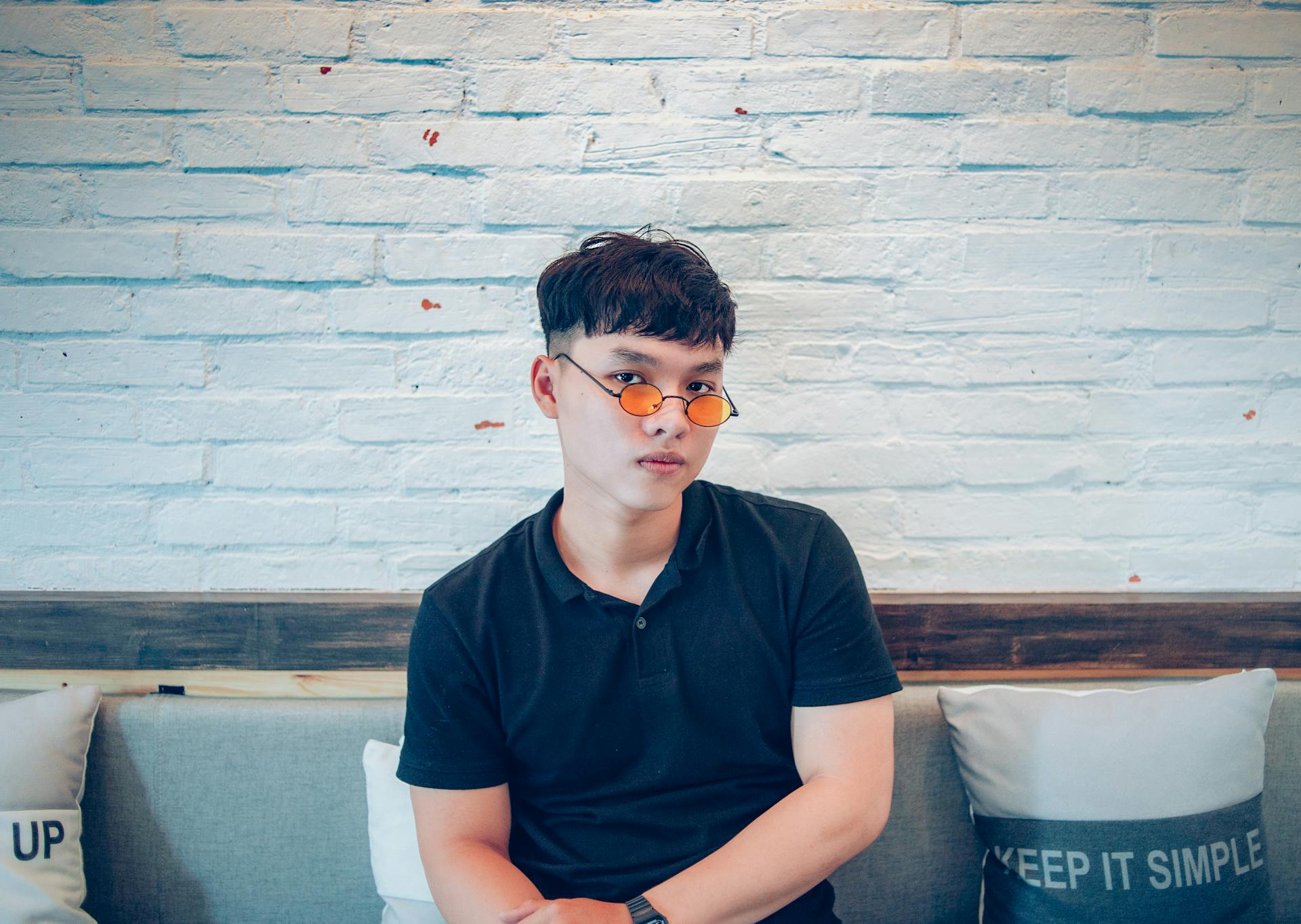 Portrait of a young Asian man sitting indoors wearing stylish sunglasses against a brick wall.