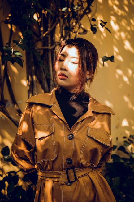 Woman in a brown coat basks in sunlight in a serene garden setting, eyes closed and relaxed.