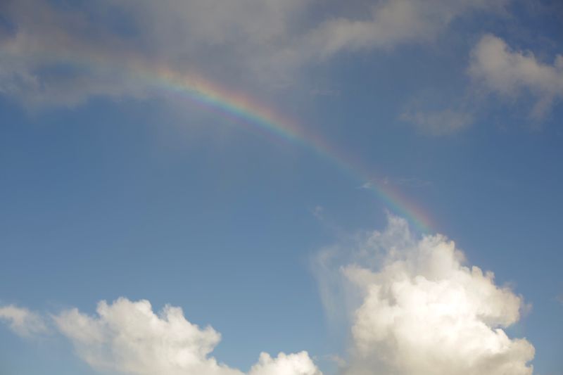 A vivid rainbow arcs over fluffy clouds against a clear blue sky.