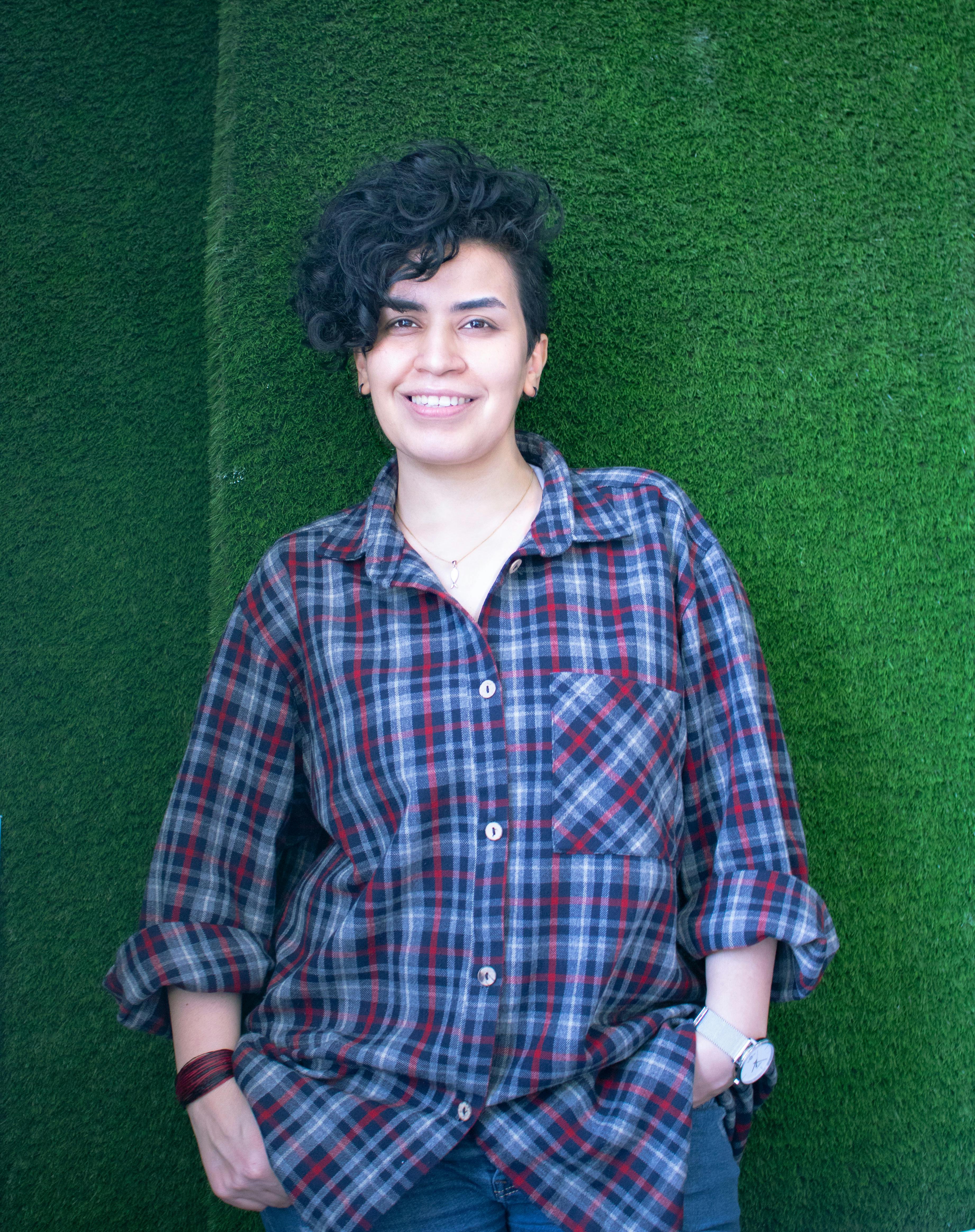 A woman with curly hair and plaid shirt smiling against a green backdrop, exuding casual confidence.