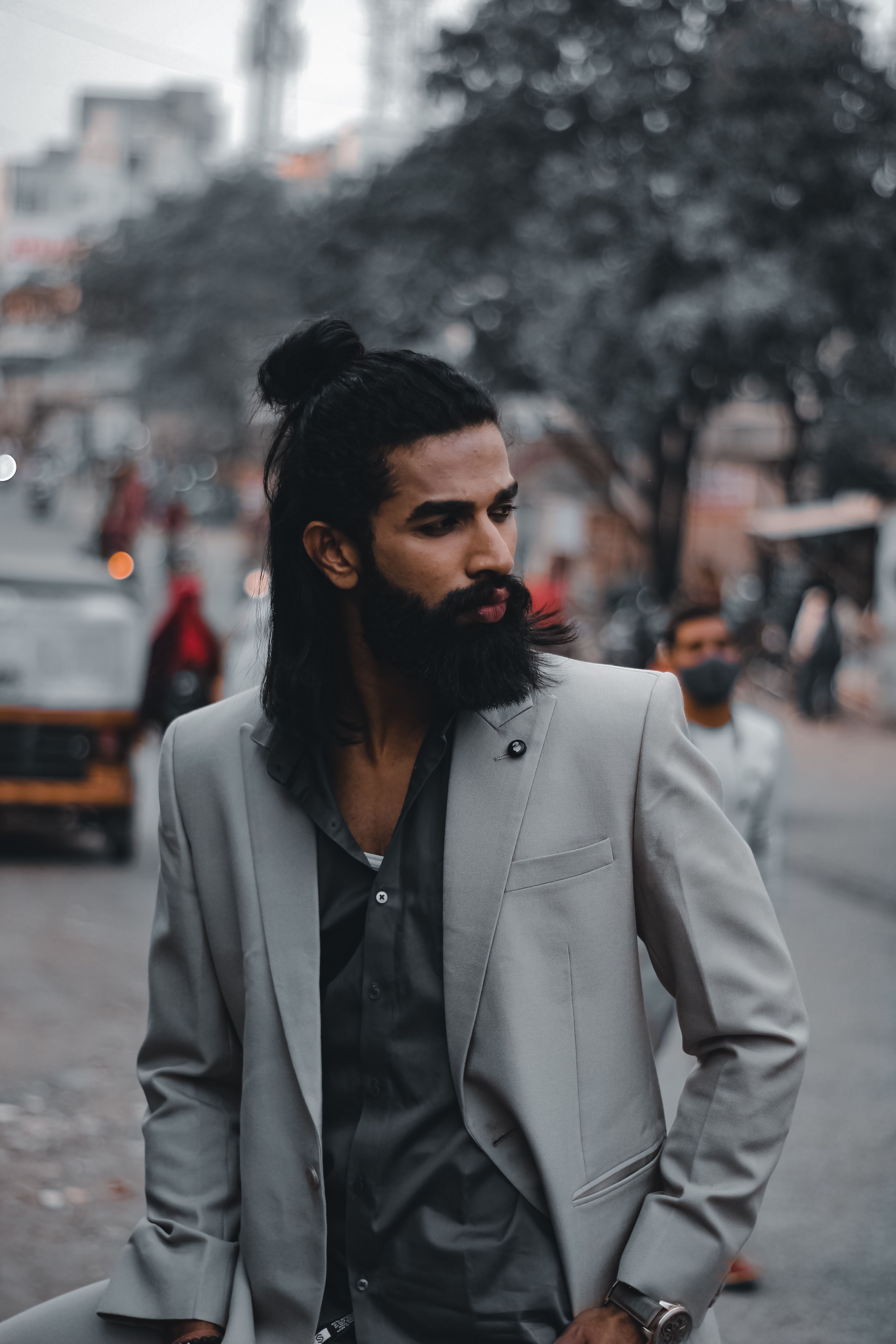 Bearded man with long hair in a gray suit posing outdoors on a busy city street.