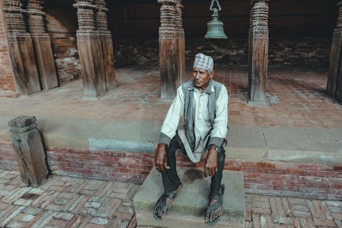 Photo of an Elderly Indian Man Sitting on Steps in front of Columns and a Brass Bell
