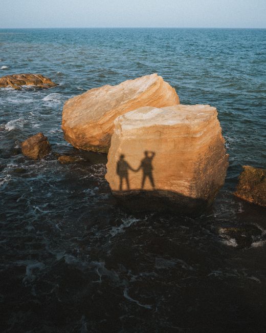 Silhouette of a couple holding hands on large coastal boulders during sunset, creating a romantic and scenic view.