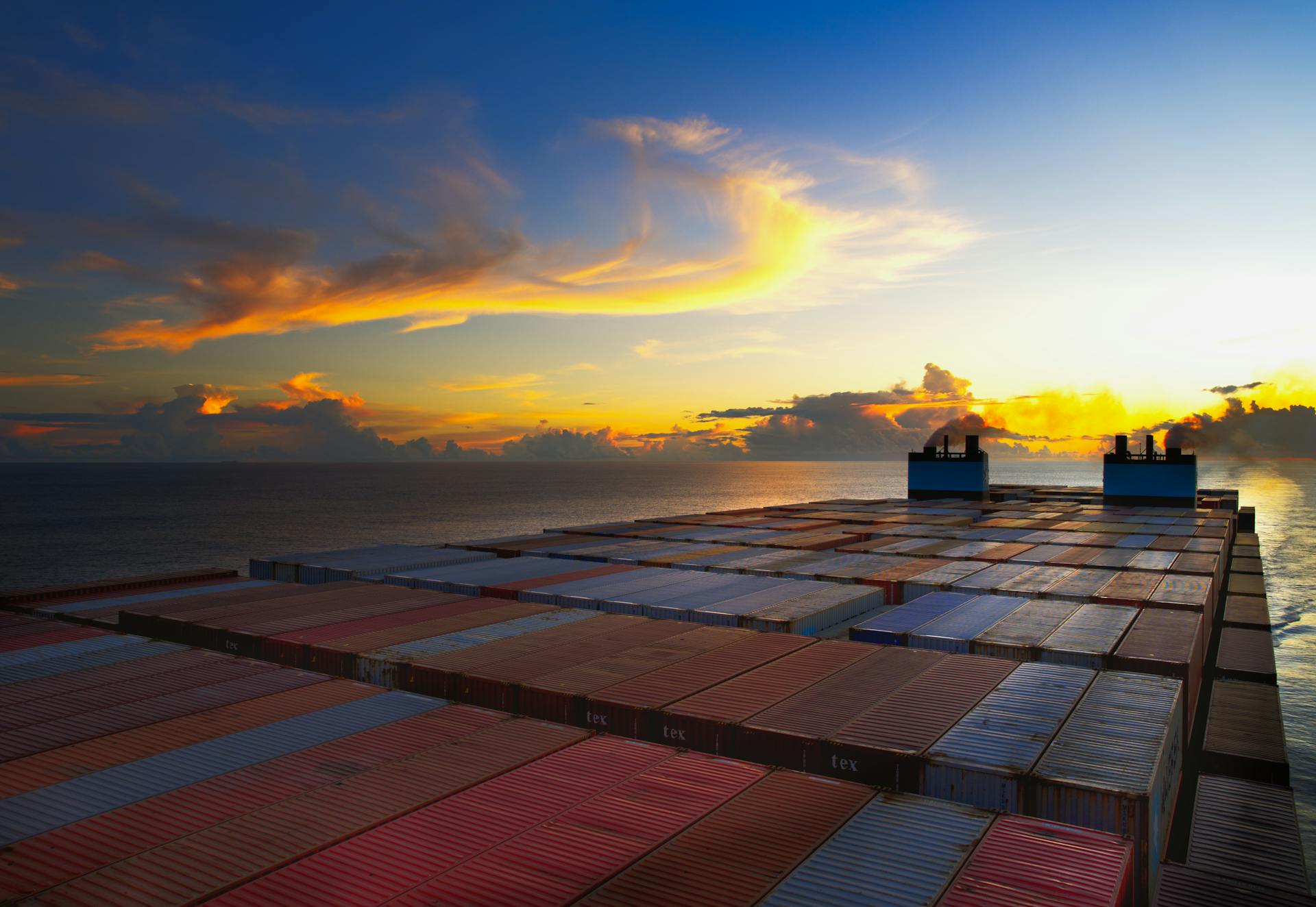 Aerial view of colorful shipping containers on a vessel against a stunning sunset sky over the ocean.