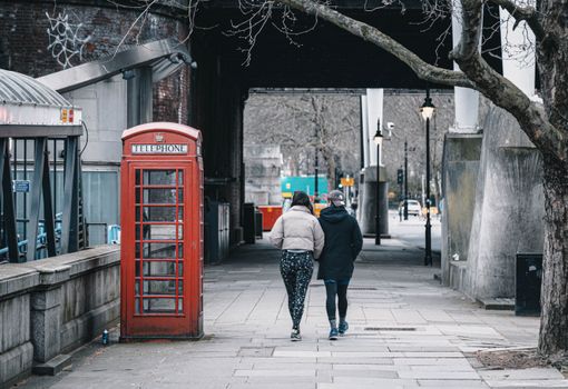A couple strolls past a classic red telephone booth in London's urban street setting.