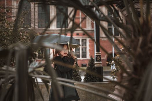 Woman in black coat holding umbrella in a lush urban London backdrop.