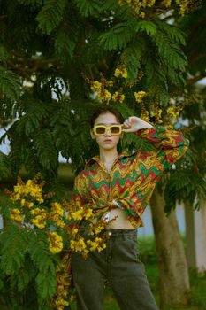 Asian woman in vibrant outfit posing with sunglasses amid lush greenery.