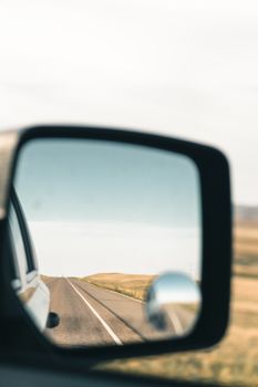 A scenic road is reflected in the side mirror of a car under a clear blue sky.