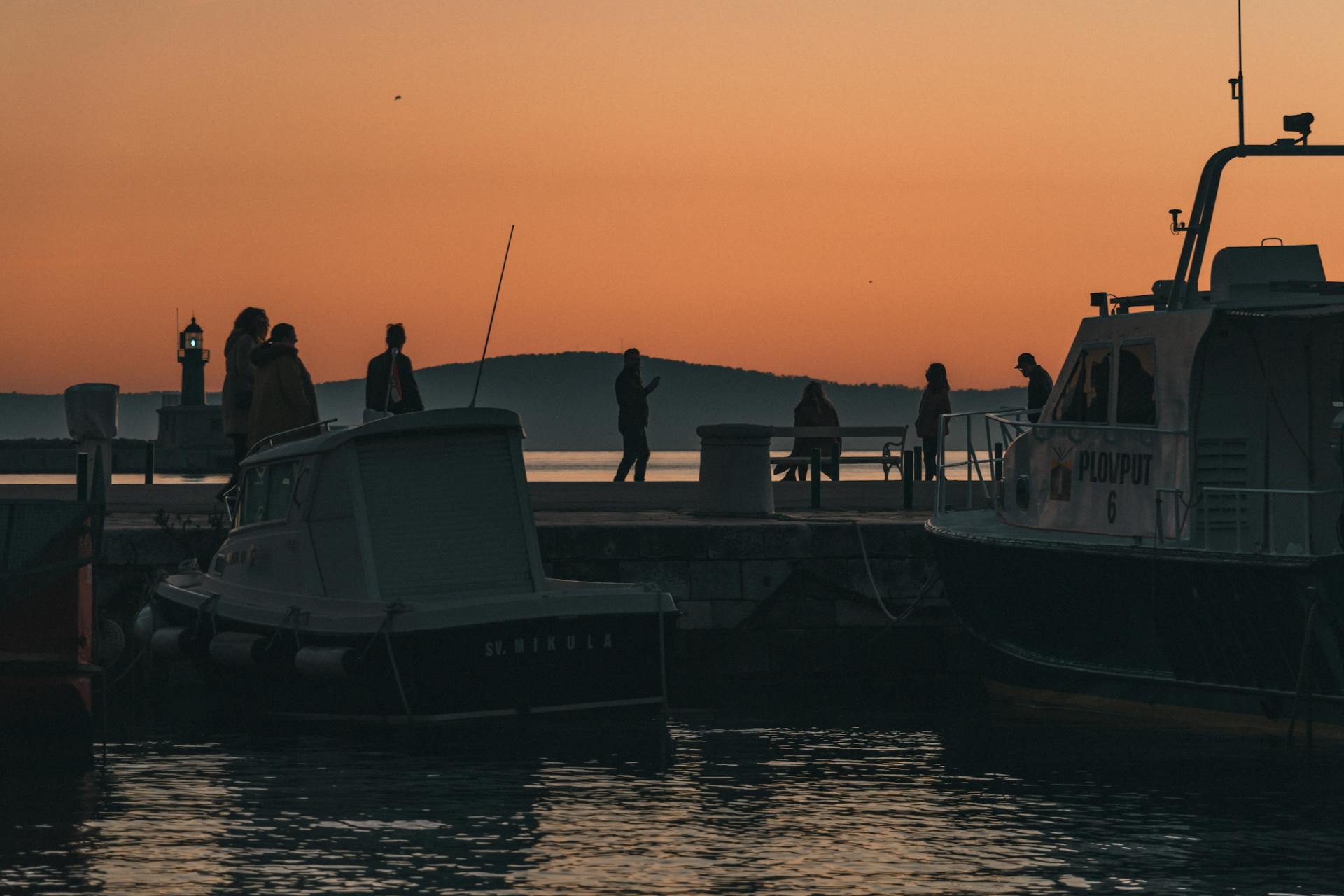 Silhouettes of people walking along the pier at sunset with boats docked at Split harbor.