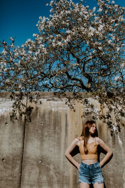 Stylish woman posing with hands on waist by a concrete wall and blossoming tree under a clear blue sky.