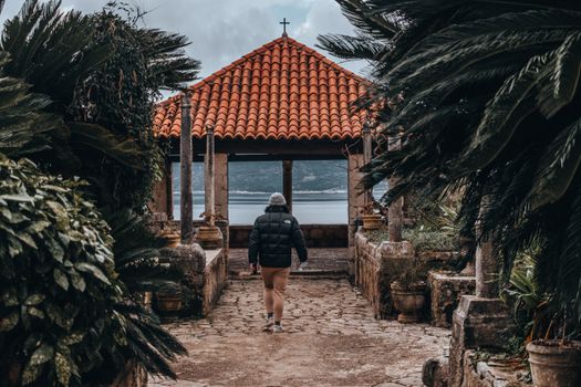 A person in a puffer jacket walks towards a gazebo overlooking the sea in a lush botanical garden.