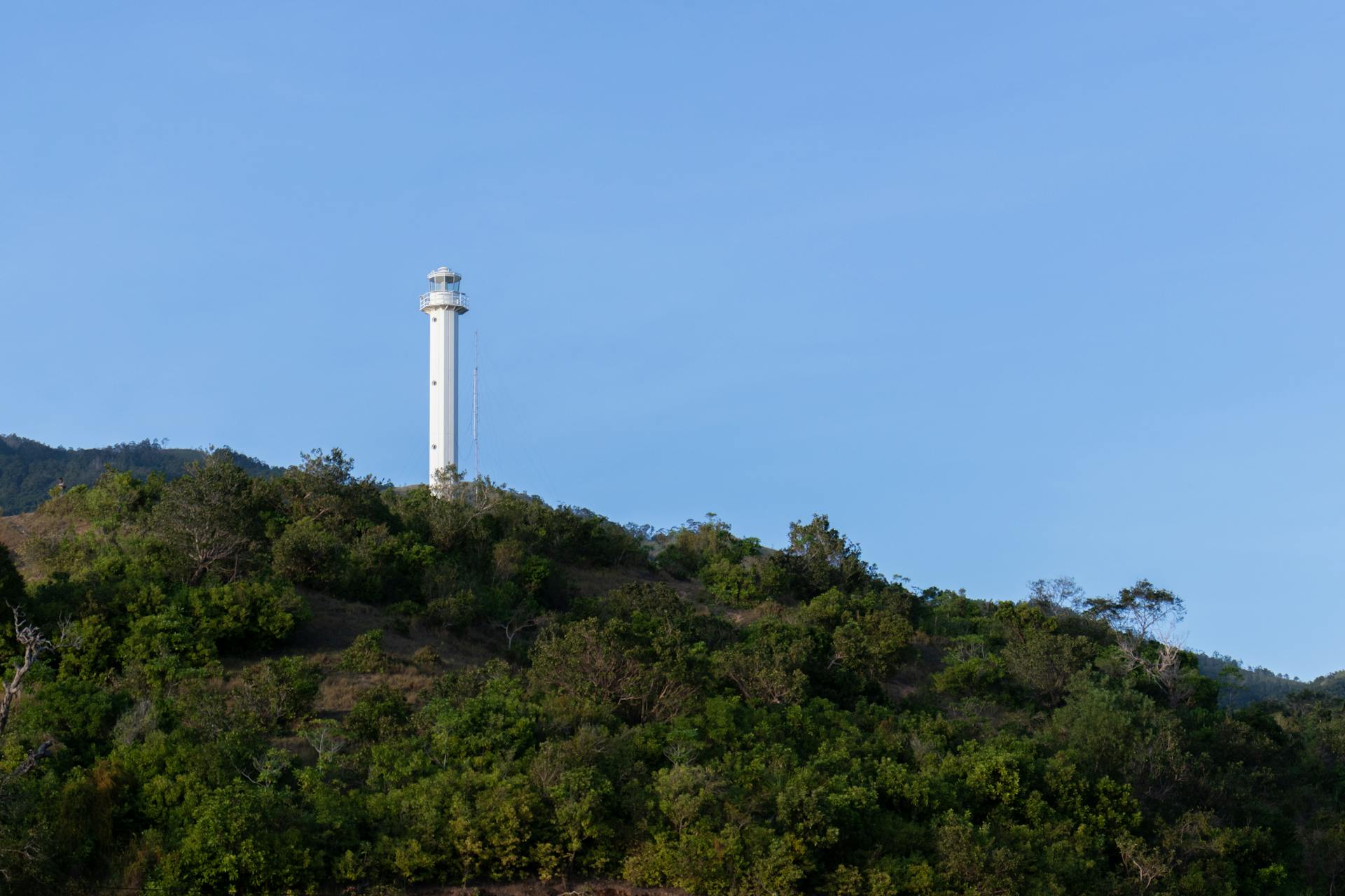 Stunning lighthouse atop lush green hill under a clear blue sky in the Philippines.