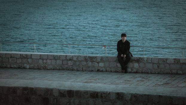 A solitary man in black clothes sits on a seawall overlooking the Adriatic sea in Dubrovnik.