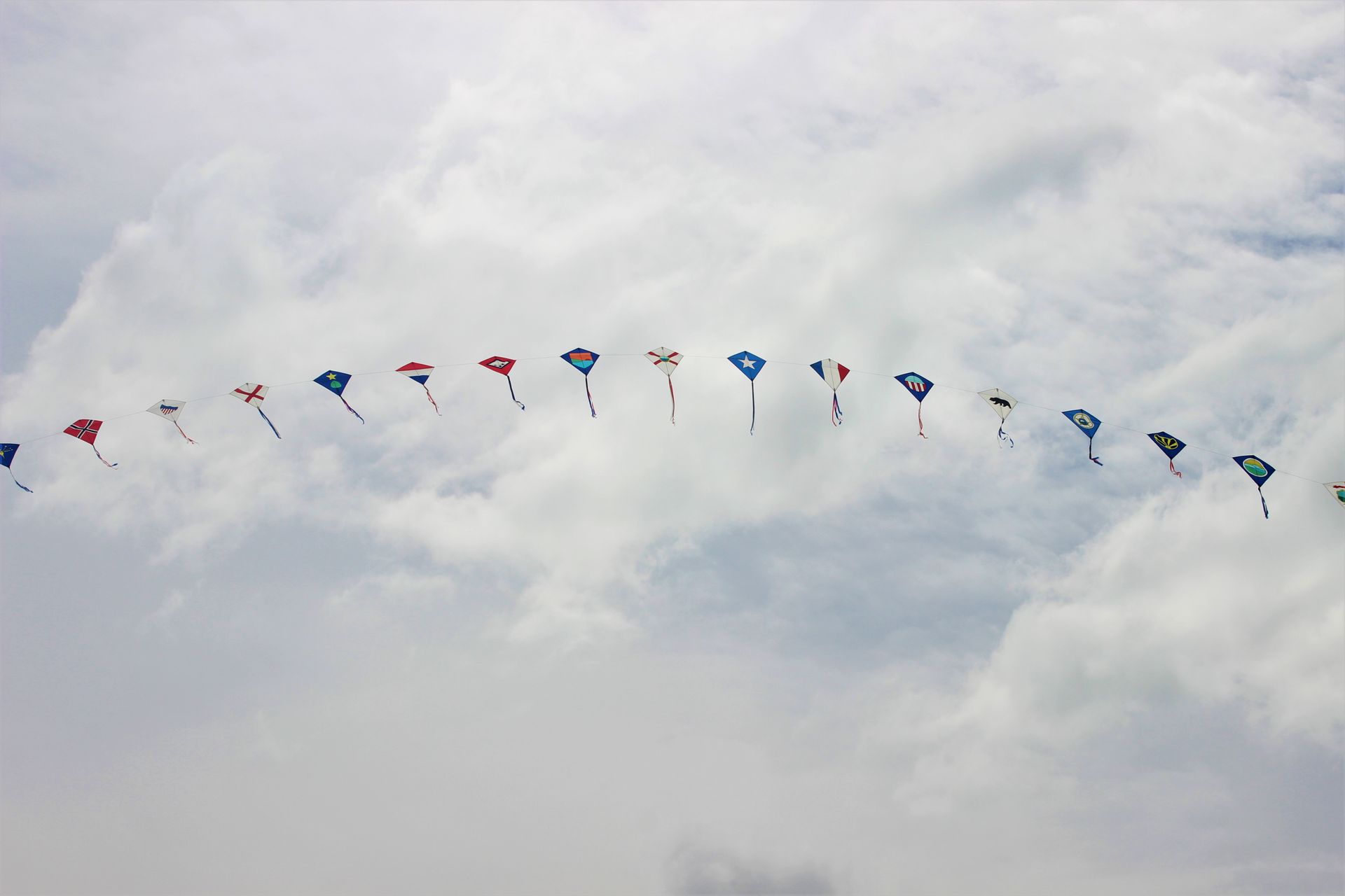 A vibrant string of kites flies under a cloudy sky, showcasing different colors and designs.