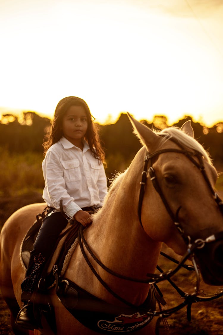 A Young Girl In White Long Sleeve Shirt Riding A Brown Horse