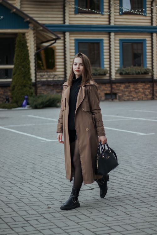 Free Stylish woman in a brown coat holding a black handbag, walking on urban cobblestone street. Stock Photo