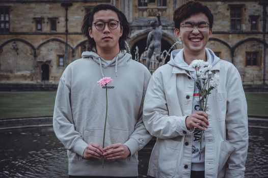 Two young men standing in a historic English courtyard, holding flowers.