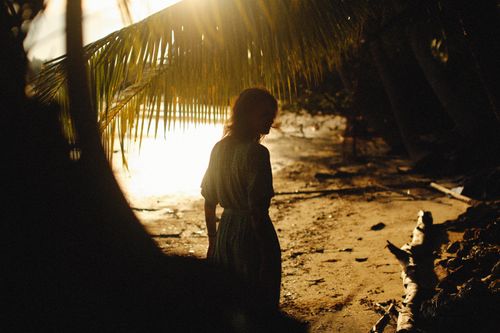 Free A woman walking on a tropical beach, silhouetted by the setting sun through palm trees. Stock Photo