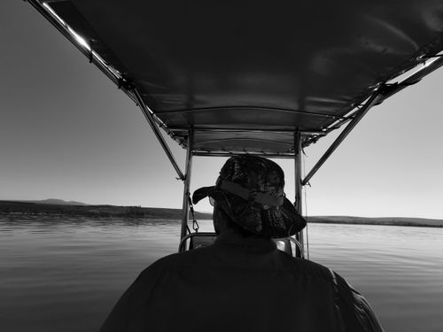 Free A man in a hat navigates a boat on a serene lake in this black and white photograph. Stock Photo