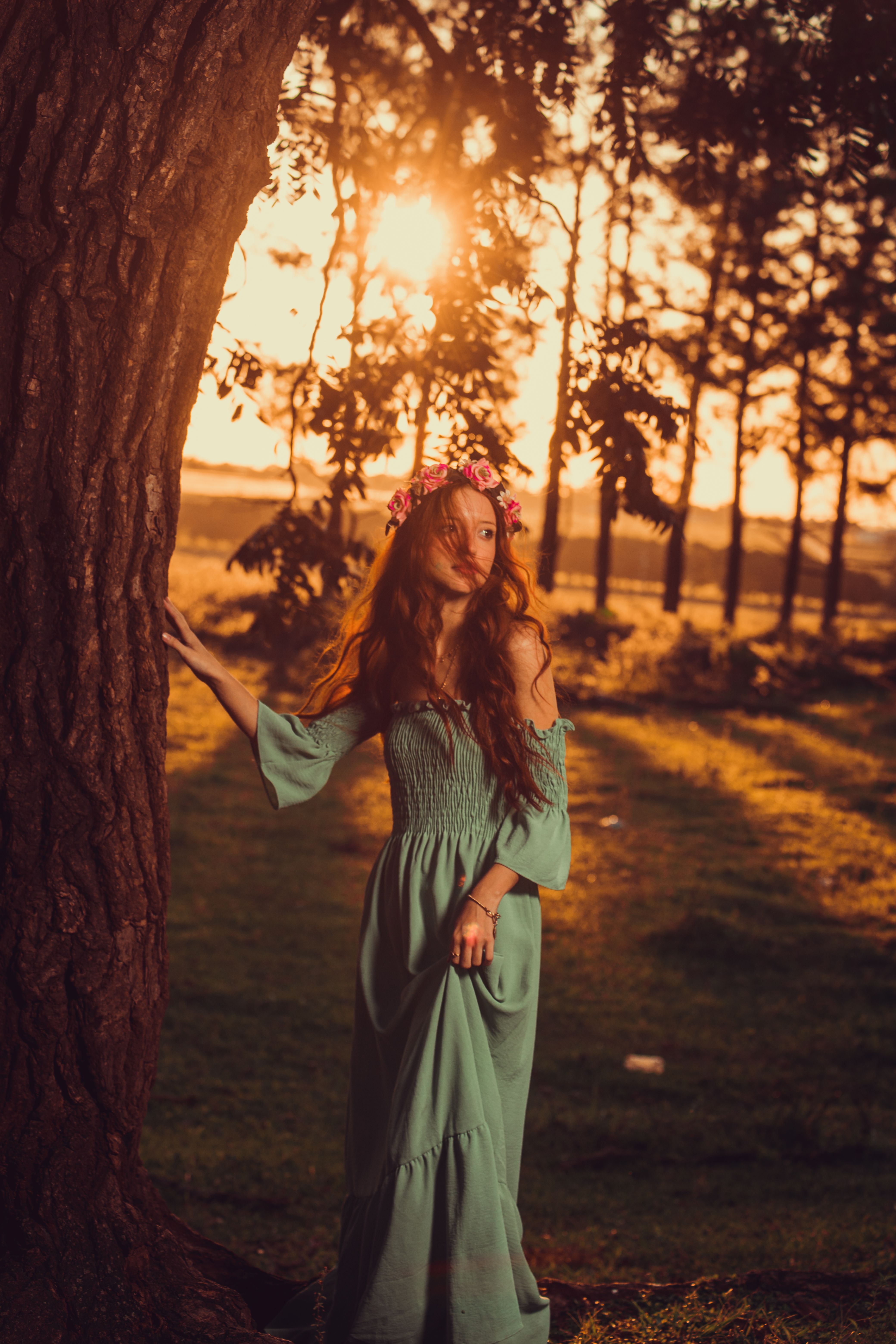 Elegant woman in a flowing dress and floral crown stands by a tree at sunset.