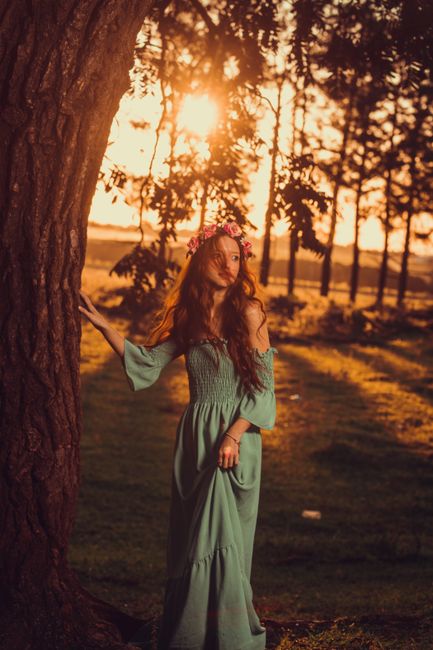 Elegant woman in a flowing dress and floral crown stands by a tree at sunset.