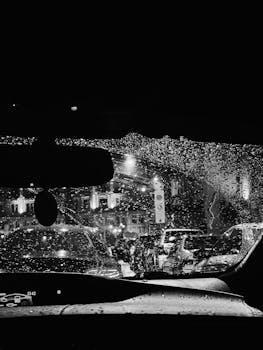 Black and white view of rainy street through a car windshield in Ukraine, featuring city lights and traffic.