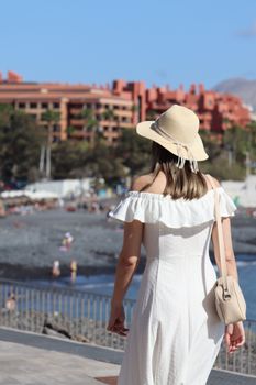 Back view of a woman in a sun hat and white dress walking along a beachfront promenade.