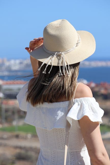 A woman in a white dress and sun hat overlooks a seaside landscape on a sunny day.