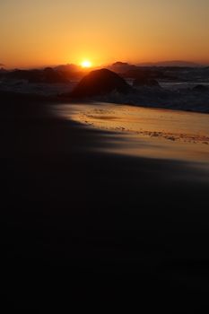 A tranquil beach scene at sunset with shimmering waves and silhouetted rocks.
