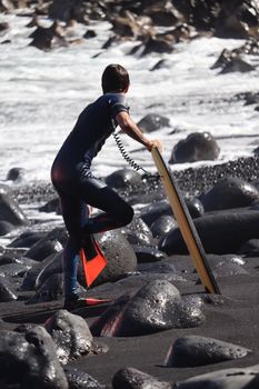 A wakeboarder in a wetsuit stands on rocks by the sea, ready for water sports.