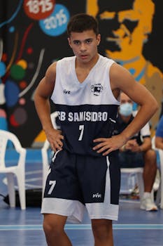Young athlete wearing CB San Isidro jersey stands focused on indoor basketball court.