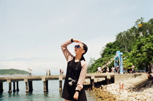 Free Asian woman enjoying a sunny day by the seaside pier, wearing sunglasses and holding a camera. Stock Photo