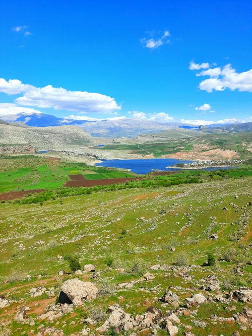 Free Breathtaking view of mountains, a serene lake, and vibrant green fields under a blue sky with white clouds. Stock Photo