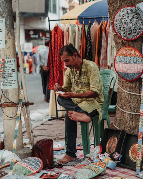 Free A male artist sitting barefoot on a street creating art amidst a vibrant market crowd. Stock Photo