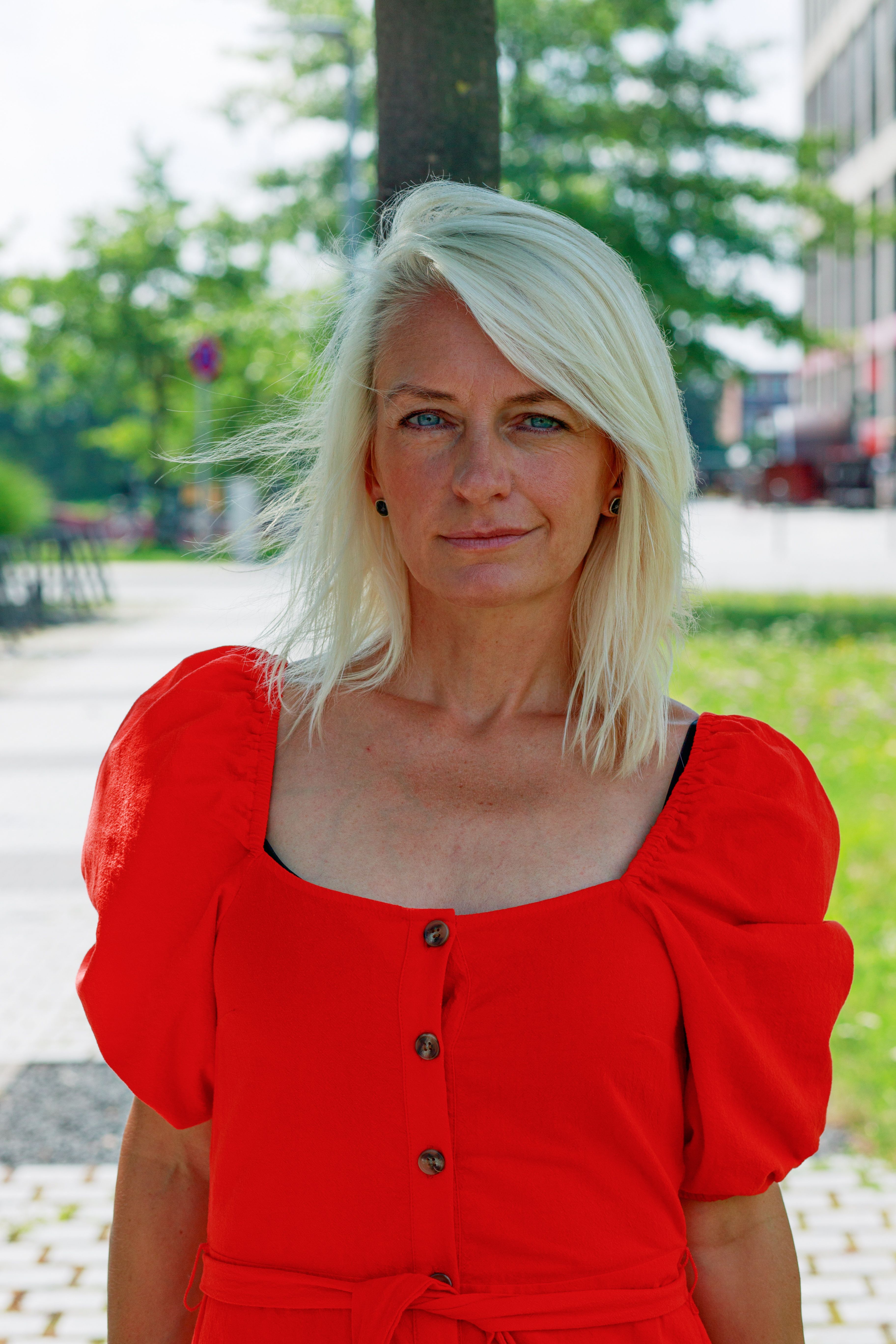 Blonde Caucasian woman in a vibrant red dress posing outdoors on a sunny day.