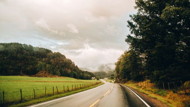 A picturesque Norwegian road traverses lush green countryside under a cloudy sky.