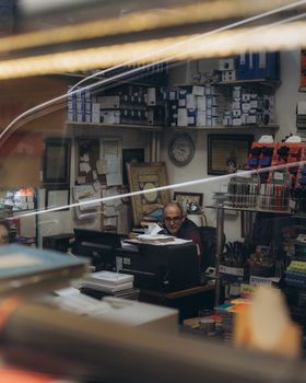 An office setting featuring a man at a desk surrounded by paperwork, office supplies, and shelves filled with files.