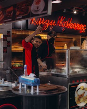 Two chefs in a street food stall preparing traditional Turkish Kuzu Kokoreç with a neon sign.