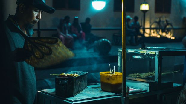Man grilling skewers at a lively night market atmosphere.