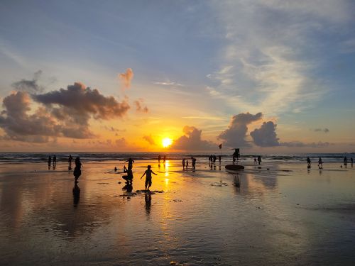 Free Silhouetted figures play at the beach during a vibrant sunset, reflecting on the wet sand. Stock Photo
