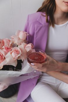 Stylish woman in a pink jacket holds a glass of rose wine and a bouquet of pink roses.