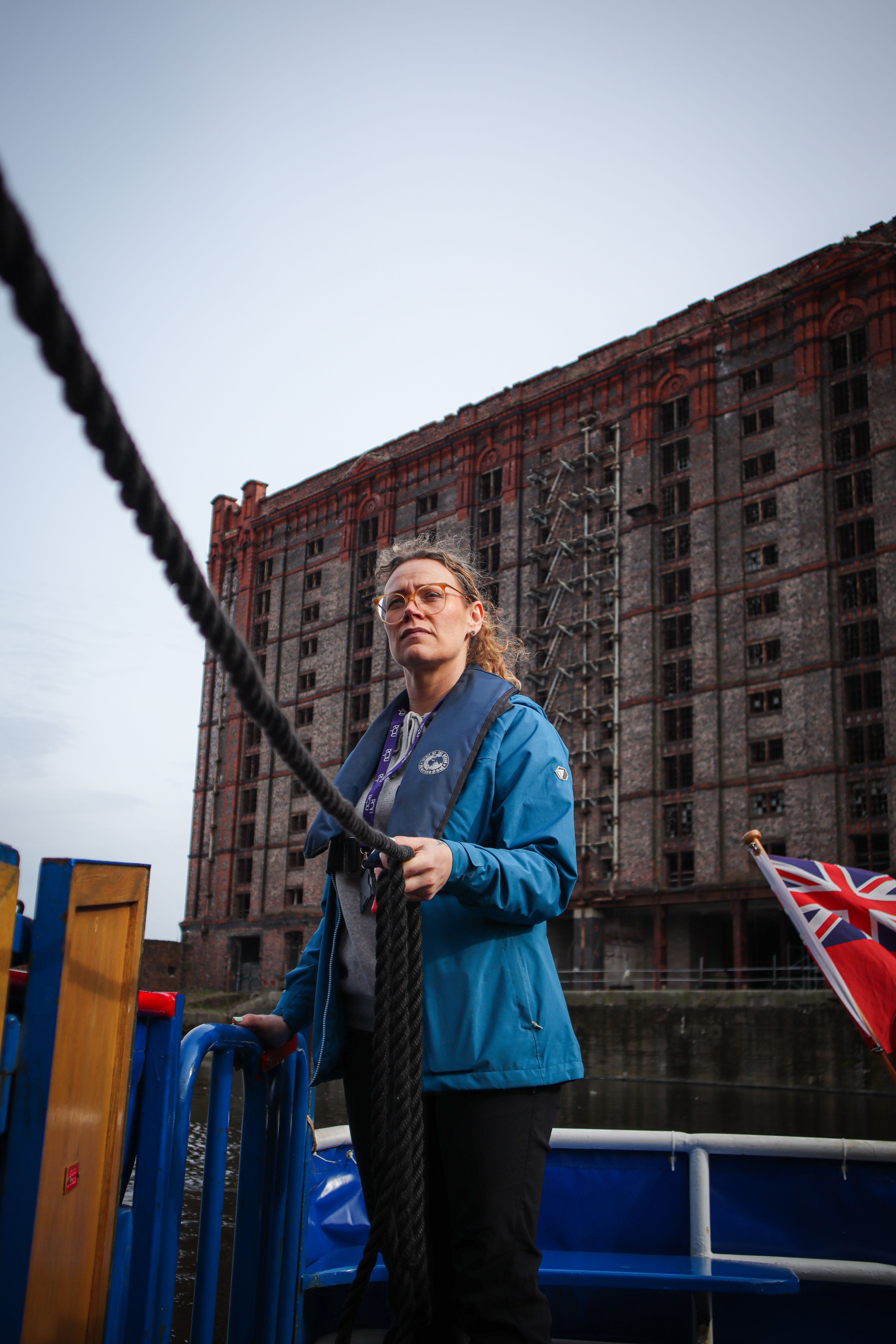 A woman in a blue jacket holds a rope on a boat in front of a historic building in England.