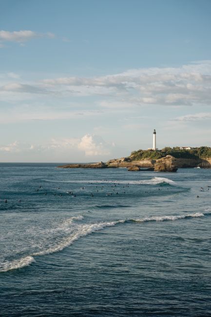 A lone lighthouse stands on a rocky shore under a clear blue sky, overlooking the sea with gentle waves.