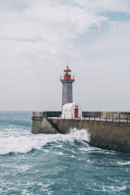 Scenic view of a lighthouse by the ocean with waves crashing against the shore under a cloudy sky.