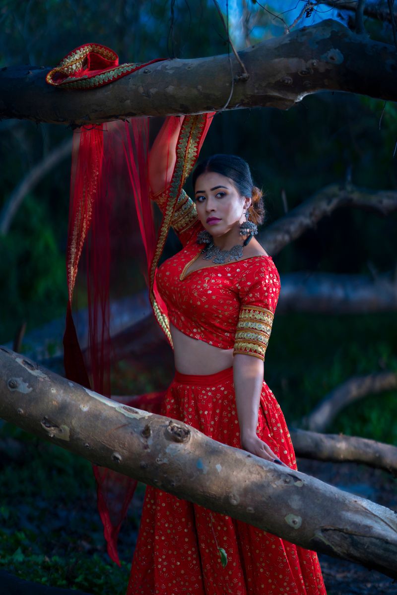 A woman in a traditional red saree poses elegantly amidst tree branches in a serene outdoor setting.