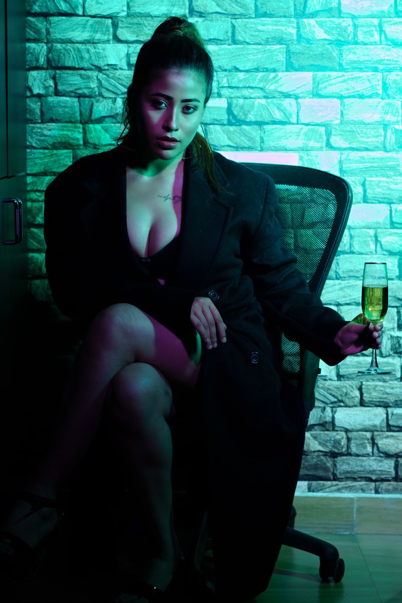 A dramatic portrait of a woman sitting indoors with a glass of wine, under moody lighting.