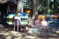 Artist painting Angkor Wat in Siem Reap, Cambodia, with vibrant canvases displayed outdoors.